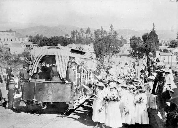 Photograph of the corner of Downey Avenue and North Broadway during the opening of the cable line to East Los Angeles, 1888. There is a cable car adorned with several American flags resting on a track at left. At right, a large crowd full of women in white dresses has gathered to watch the ceremony. Down the street, more people have gathered on the sidewalks. There are several large buildings visible, as well as a large number of trees. The cable car bears the number 60.